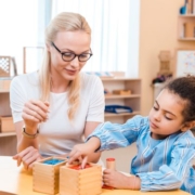A képen egy szemüveget pedagógus nézi ahogy egy kislány dobozokba színek szerint válogatja ki a játékokat