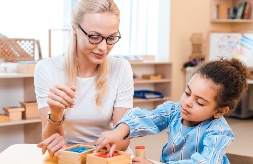A képen egy szemüveget pedagógus nézi ahogy egy kislány dobozokba színek szerint válogatja ki a játékokat