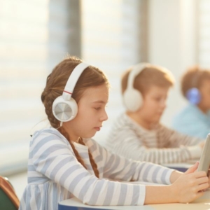 Gyerekek tablettel a kézben, fejhallgatóval a fejükön ülnek egy teremben egymás mellett.
