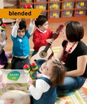 A gyermekek szükségleteire épülő mindennapokban az éneklés, zenélés támogató jelenléte kiemelt kép, amelyen feltüntettük hogy blended típusú továbbképzésről van szó. A kiemelt fotón gyerekek énekelnek zenélnek egy pedagógus körül