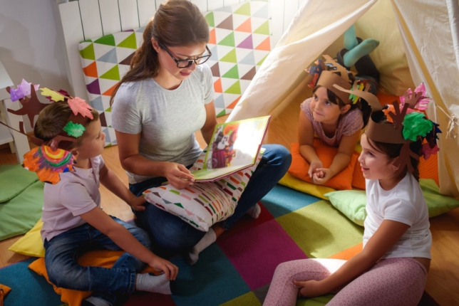 A képen egy szemüveges nő olvas mesét három kisgyereknek, akik színes fejdíszeket viselnek. Az egyikük beltéri játszó sátorban hason fekszik, a másik két gyerek ül.