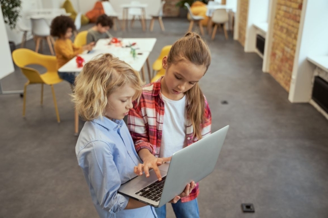 Egy kényelmes, otthonos teremben két gyerek áll, kezükben egy laptop. A háttérben egy asztalnál másik két gyerek ül.