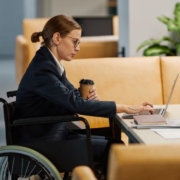 Rehabilitációs hozzájárulás kiemelt kép. Egy csinosan öltözött szemüveges hölgy ül egy elegáns kanapékkal körülvett dohányzóasztalnál. A laptopján dolgozik, az egyik kezében kávés poharat tart.