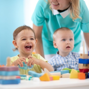 A képen mosolygó kisgyermekek játszanak az asztalon építőkockákkal, a háttérben mosolygó kisgyermeknevelő áll mögöttük