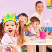 Gyerekek ülnek az óvodában, előtérben egy kislány, fején egy tolldísz, az óvónénit a háttérben látjuk