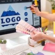 A bölcsőde logója kiemelt kép - a képen egy pantone színskálát tart egy női kéz, a háttérben monitoron egy hegyet ábrázoló logo terv