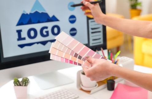 A bölcsőde logója kiemelt kép - a képen egy pantone színskálát tart egy női kéz, a háttérben monitoron egy hegyet ábrázoló logo terv