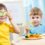 A képen két kisfiú ül az asztalnál, egyikük sárga paprika karikákat fog a kezében, a másik két szelet uborkát tart a szemei elé. Az asztalon egy tányéron sok zöldség és gyümölcs található