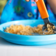 A képen egy kisgyermek kék tányérja látható, benne rizsszemek és egy narancssárga markolatú evőeszköz. A háttérben csak egy kék színű partedli látszik.