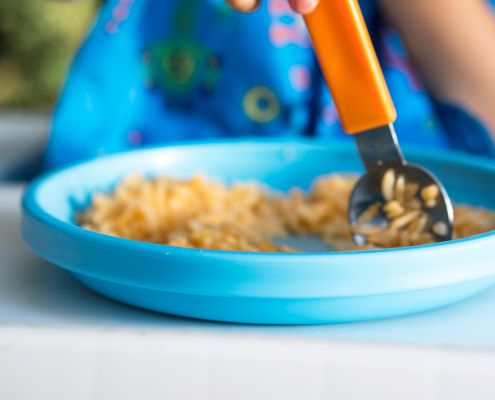 A képen egy kisgyermek kék tányérja látható, benne rizsszemek és egy narancssárga markolatú evőeszköz. A háttérben csak egy kék színű partedli látszik.