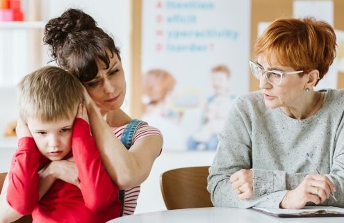 A képen egy középkorú, szemüveges nő füzettel és tollal a kezében néz egy édesanyát, aki az ölében ülő, füleit befogó kisfiú fejét fogja, és a fejét a kisfiú fejéhez érinti gyengéden, együttérzéssel.