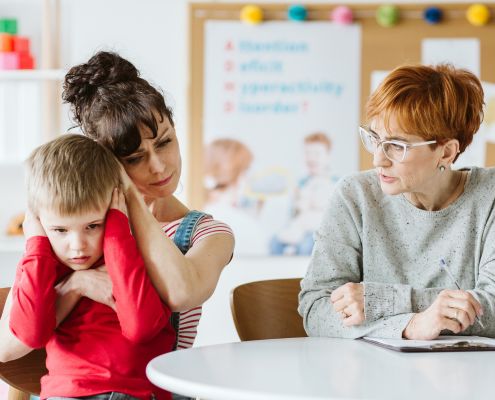 A képen egy középkorú, szemüveges nő füzettel és tollal a kezében néz egy édesanyát, aki az ölében ülő, füleit befogó kisfiú fejét fogja, és a fejét a kisfiú fejéhez érinti gyengéden, együttérzéssel.