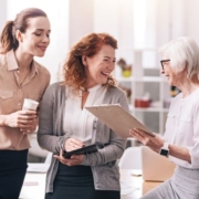 A képen egy irodában három eltérő korú nő beszélget, nevet