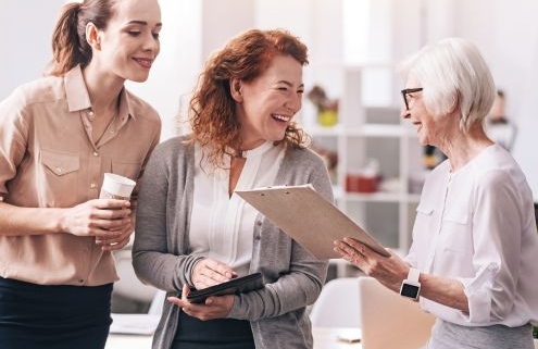 A képen egy irodában három eltérő korú nő beszélget, nevet