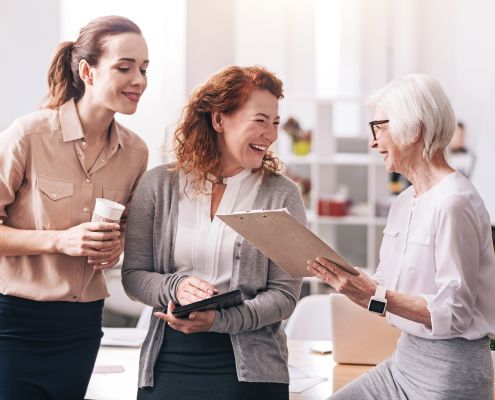 A képen egy irodában három eltérő korú nő beszélget, nevet