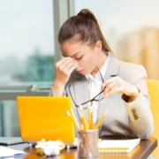 Elég az egészből, ég veled, kiégés! Egy fiatal nő ül a sárga laptopja mögött, kezét a homloka közepére helyezve. Másik kezében a szemüvegét tartja. A ceruzatartóban a laptoppal egyező színű ceruzák vannak