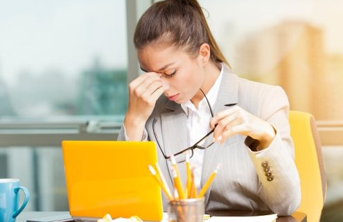 Egy fiatal nő ül a sárga laptopja mögött, kezét a homloka közepére helyezve. Másik kezében a szemüvegét tartja. A ceruzatartóban a laptoppal egyező színű ceruzák vannak