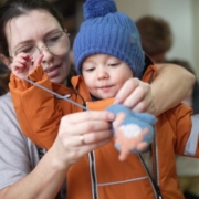 Az önálló öltözködés játékos támogatása - kisgyermeknevelő segíti a kesztyű felvételét egy anorákba bújt kisfiúnak