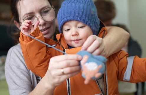 Az önálló öltözködés játékos támogatása - kisgyermeknevelő segíti a kesztyű felvételét egy anorákba bújt kisfiúnak