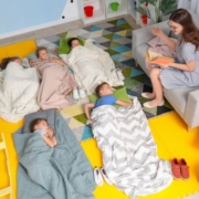 Kisgyermekek fekszenek a földön az óvodában vagy bölcsődében, a pedagógus a kanapén ül, mosolyogva mesét olvas nekik.