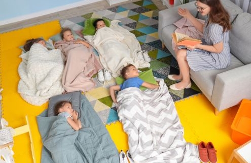 Kisgyermekek fekszenek a földön az óvodában vagy bölcsődében, a pedagógus a kanapén ül, mosolyogva mesét olvas nekik.