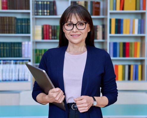 Márciusi óvodavezetői feladatok - a képen egy szemüveges középkorú, csinosan öltözött nő áll, kezében egy laptop, mögötte könyvespolcok színes könyvekkel.