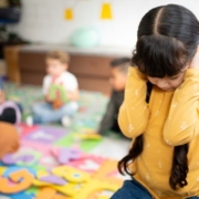 Szenzorosan érzékeny gyerekek az óvodában A képen egy sárga pulcsis, sötét hajú kislány az óvodai csoportszobában a kezeit a fülére tapasztva, egyedül térdel