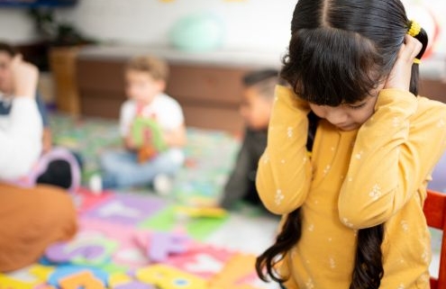 A képen egy sárga pulcsis, sötét hajú kislány az óvodai csoportszobában a kezeit a fülére tapasztva, egyedül térdel