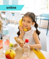 Egy kislány az óvodai csoportszobában egy szendvicset eszik, a másik kezében egy banán, amit az arcához szorít