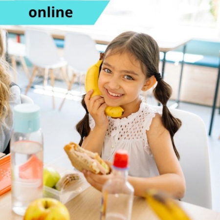 Egy kislány az óvodai csoportszobában egy szendvicset eszik, a másik kezében egy banán, amit az arcához szorít