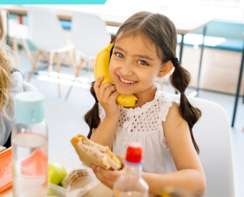 Egy kislány az óvodai csoportszobában egy szendvicset eszik, a másik kezében egy banán, amit az arcához szorít
