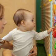 A kisgyermeknevelők mozgásszervi problémái - egy kisgyermeknevelő egy kisbabát tart az ölében, feltehetően guggolva, a baba a falra szerelt játékot babrálja