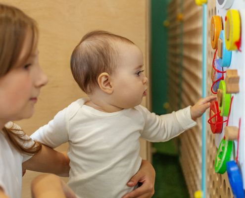 A kisgyermeknevelők mozgásszervi problémái - egy kisgyermeknevelő egy kisbabát tart az ölében, feltehetően guggolva, a baba a falra szerelt játékot babrálja