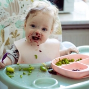 Csecsemőétkeztetés a bölcsődében - egy maszatos kisbaba etetőszékben ül, kezében kiskanál, előtte egy rózsaszín tányér, és az etetőszék tele ételdarabkákkal