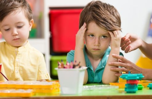 Dühös kisfiú ül az óvodai asztalnál a társa mellett. Egy óvónő simogatja a karját, próbálja megnyugtatni