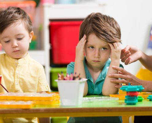 Dühös kisfiú ül az óvodai asztalnál a társa mellett. Egy óvónő simogatja a karját, próbálja megnyugtatni