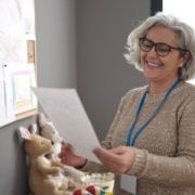 Egy idősebb óvónő egy lapot tart a kezében egy gyerekrajzokat tartalmazó tábla előtt.