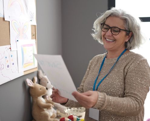 Egy idősebb óvónő egy lapot tart a kezében egy gyerekrajzokat tartalmazó tábla előtt.