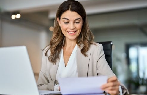 Egy kedves arcú nő ül a laptop előtt, kezében iratok, azokat nézi át