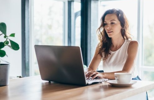 Hogyan készüljünk a minősítési eljárásra - egy fiatal nő otthon, egy fényes lakásban az asztalnál ül a laptopja előtt