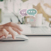 Online kommunikáció a bölcsődevezetésben A képen egy női kéz egy egér fölött. A levegőben két beszédbuborék grafika található, a buborékokban három három pont. A háttérben egy fehér laptop és egy kávés bögre