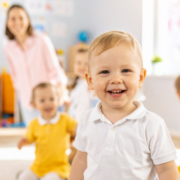 Kisgyermekek önállóságának segítése - a képen egy bölcsődés kisfiú mosolyog az előtérben. A háttérben egy másik totyogó, valamint a kisgyermeknevelő látszik elmosódva