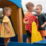 Az óvodai konfliktusok kezelése Az óvodai konfliktusok kezelése - az óvodai játszótéren kés kisfiú összeszólalkozik a mászóka tetején, egy kormány előtt. Egy sárga ruhás kislány nézi őket.