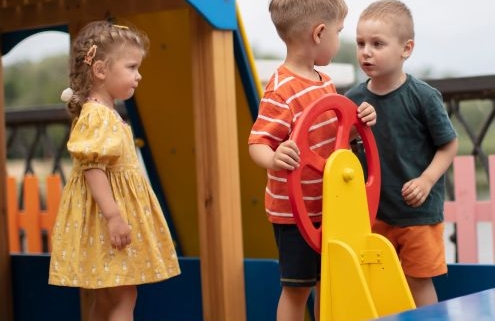 Az óvodai konfliktusok kezelése - az óvodai játszótéren kés kisfiú összeszólalkozik a mászóka tetején, egy kormány előtt. Egy sárga ruhás kislány nézi őket.