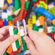 LEGO-val az iskolába – Játéklehetőségek korosztályonként Egy kisgyermek kezét látjuk aki LEGO-ból épít valamit. A háttérben is LEGO kockák vannak
