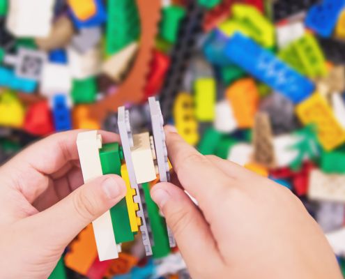Egy kisgyermek kezét látjuk aki LEGO-ból épít valamit. A háttérben is LEGO kockák vannak